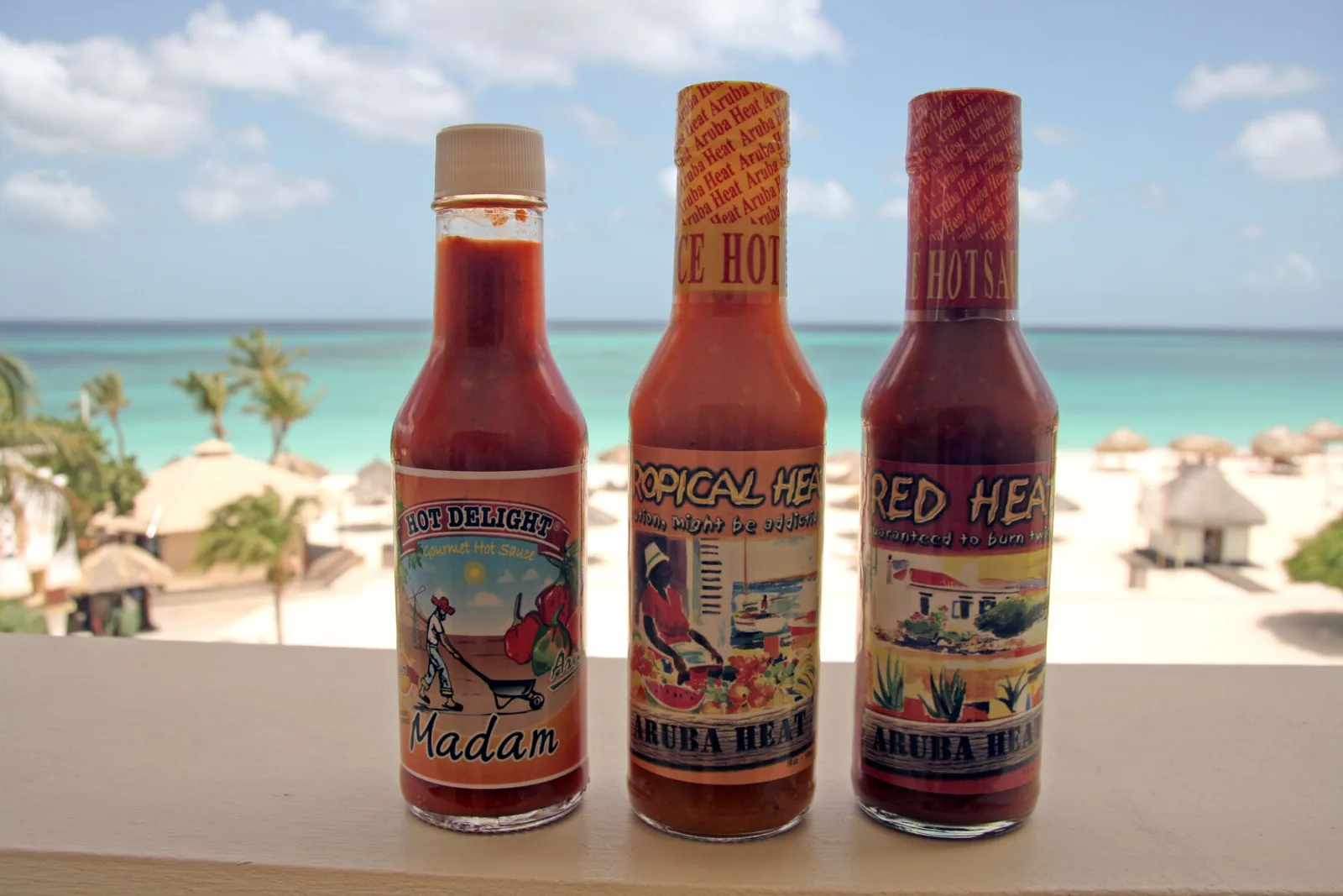 Three bottles of Aruban hot sauce, Hot Delight Madam and Aruba Heat's Tropical Heat and Red Heat, on a balcony railing above a beach with palapas in Aruba
