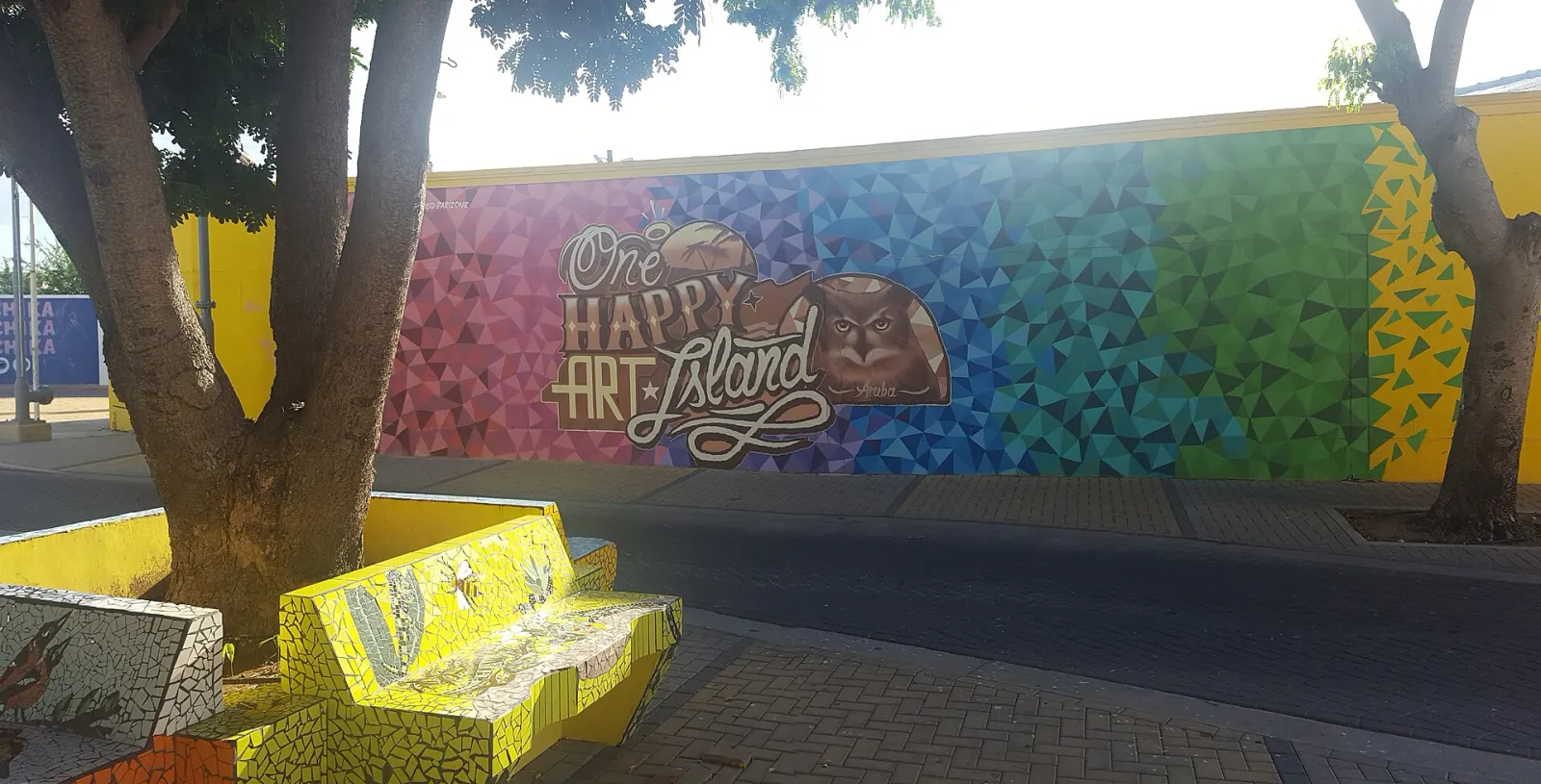 The colorful One Happy Art Island mural with a shoco owl on Zeppenfeldstraat in San Nicolas, Aruba, behind a mosaic-tiled bench