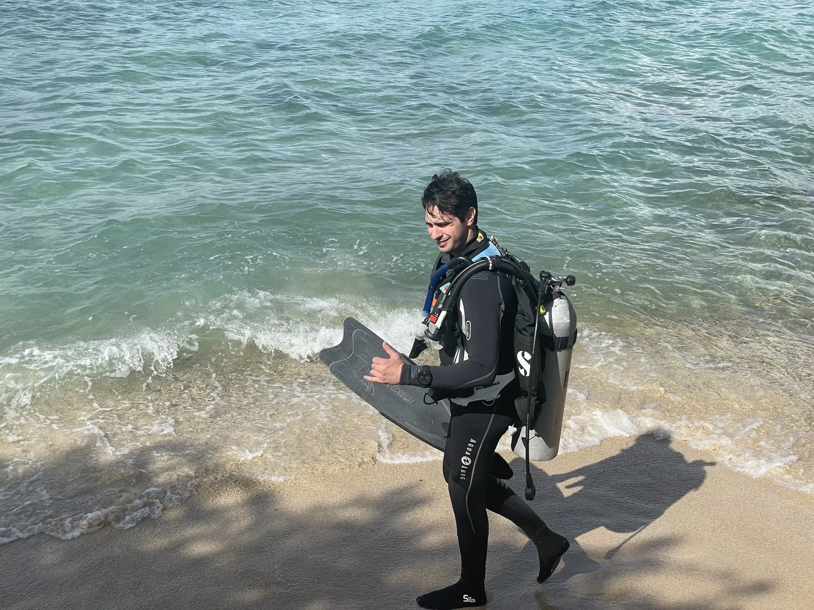 A smiling scuba diver in full gear walking out of the water onto the beach at Peachy Beach in Savaneta, Aruba, after a shore dive