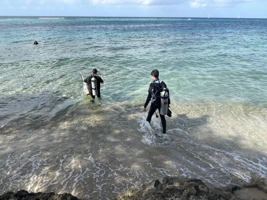 Two scuba divers walking into the water from the beach at Peachy Beach in Savaneta, Aruba, for a shore dive on the house reef, with the reef's line of breaking waves offshore
